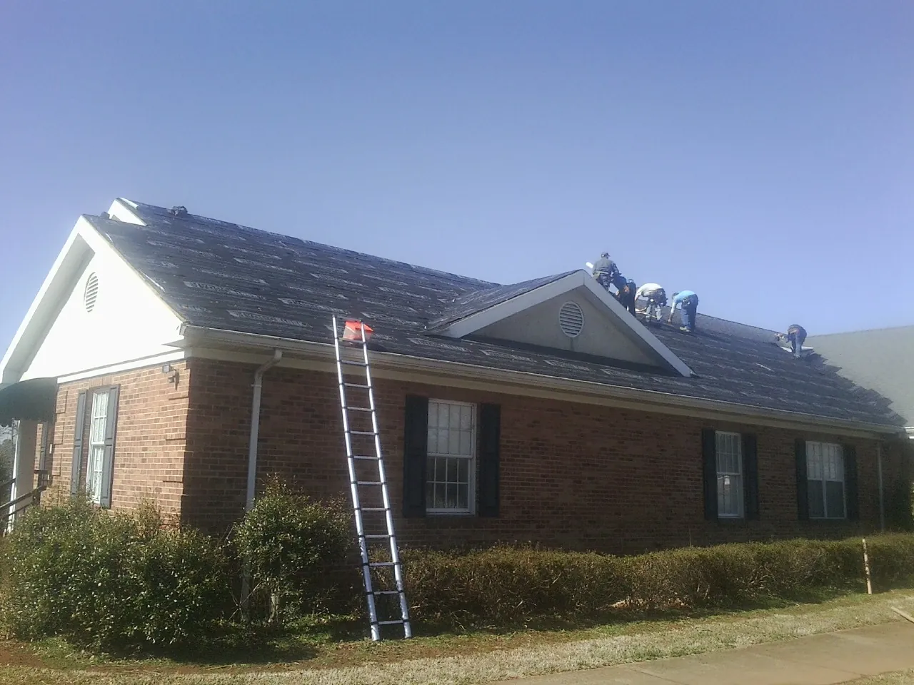 Crew tearing shingles off a brick ranch roof with a ladder set — Emergency Roof Repair work by our crew in Adrian